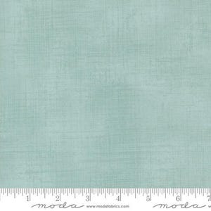 May include: A light seafoam green fabric with a subtle crosshatch texture. A white ruler is visible at the bottom edge, showing measurements in inches from 1 to 7. The text "moda" and "www.modafabrics.com" is printed along the bottom edge.