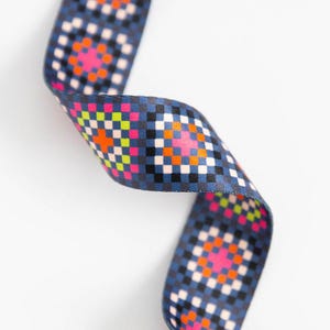 May include: A blue ribbon with a repeating geometric pattern of squares in pink, orange, green, black, and white. The ribbon is curved and appears to be made of a woven material.