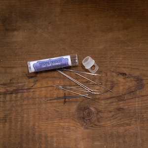 May include: A clear plastic tube containing a set of small, silver knitting needles. The tube has a label that reads "Knitting Needles" with a purple and white design. The needles are scattered on a wooden surface.