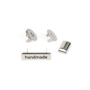 May include: Silver metal hardware set for attaching a label to a garment or other item. The set includes two snap fasteners, a rectangular metal plate with the word "handmade" engraved on it, and a separate rectangular metal plate.
