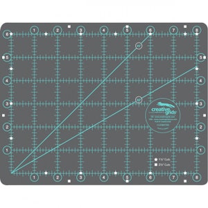 May include: A gray cutting mat with a grid pattern and teal markings for angles and measurements. The mat includes the text "creative grids" and is designed for quilting and crafting projects. It has measurements in inches.