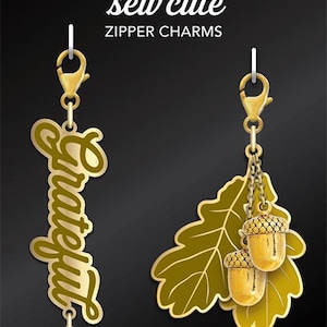May include: Two gold zipper charms, one with the word "Grateful" and the other with an oak leaf and two acorns. The charms are on a black background with the text "Cathe Holden's sew cute zipper charms" and "moda fabrics + supplies".