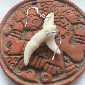 May include: A pair of earrings made from white bone, each with a silver hook. The earrings are displayed on a brown ceramic tile with a decorative design.