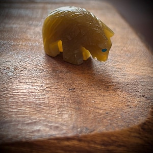 Puede incluir: Una pequeña figura de oso tallada en una piedra translúcida de color marrón dorado. El oso está representado en posición de pie con la cabeza inclinada hacia abajo. Presenta una única incrustación circular pequeña de color azul brillante.