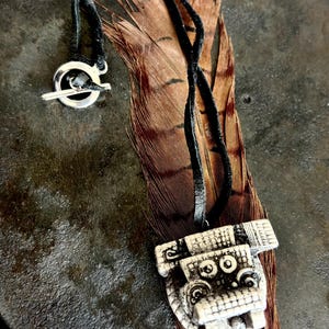May include: A pendant necklace with a black cord and a silver clasp. The pendant is a carved, off-white, rectangular shape with a detailed design. The necklace rests on a brown and black feather.