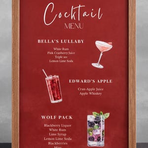 May include: A framed cocktail menu with a red background and a wooden frame. The menu features three cocktail illustrations: Bella's Lullaby, Edward's Apple, and Wolf Pack. The menu lists the ingredients for each cocktail in white text.