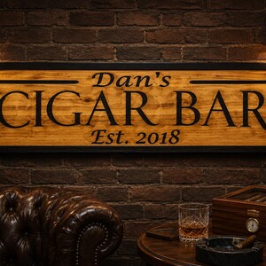 May include: Wooden sign with the text "Dan's CIGAR BAR Est. 2018" in black lettering. The sign is framed in black and mounted on a brick wall. The scene includes a leather couch, a cigar box, and a glass of whiskey.