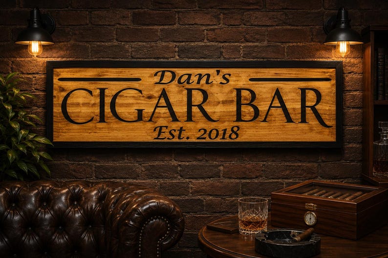 May include: Wooden sign with the text "Dan's CIGAR BAR Est. 2018" in black lettering. The sign is framed in black and mounted on a brick wall. The scene includes a leather couch, a cigar box, and a glass of whiskey.