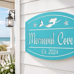 May include: A rectangular turquoise sign with rounded corners featuring a white cutout of a mermaid flanked by two starfish. The text reads Mermaid Cove in a script font above the text Est. 2024 in a smaller font. The sign has a white border.