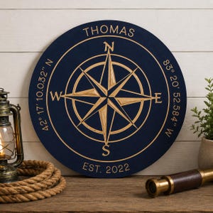 May include: A navy blue wooden compass rose with the name "THOMAS" at the top and "EST. 2022" at the bottom. The compass rose is gold-colored and features directional markings and coordinates.