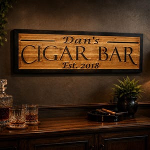 May include: Wooden sign with the text "Dan's CIGAR BAR Est. 2018" in a dark room. The sign is framed in black and mounted on a dark brown wall. A liquor cabinet, glasses, and a decanter are visible.
