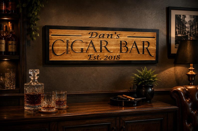 May include: Wooden sign with the text "Dan's CIGAR BAR Est. 2018" in a dark room. The sign is framed in black and mounted on a dark brown wall. A liquor cabinet, glasses, and a decanter are visible.