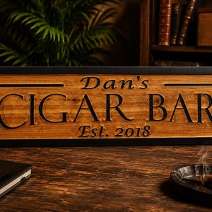May include: A wooden sign with the words "Dan's CIGAR BAR" and "Est. 2018" carved into it. The sign is framed in black and sits on a wooden surface. A lit cigar rests in a small ashtray.