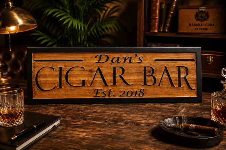 May include: A wooden sign with the words "Dan's CIGAR BAR" and "Est. 2018" carved into it. The sign is framed in black and sits on a wooden surface. A lit cigar rests in a small ashtray.