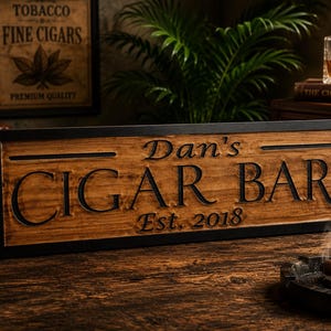 May include: Wooden sign with the text "Dan's CIGAR BAR Est. 2018" in a black frame. A lit cigar rests in a black ashtray. A glass of amber liquid and a vintage lamp are also visible.