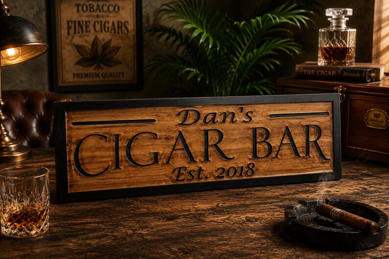 May include: Wooden sign with the text "Dan's CIGAR BAR Est. 2018" in a black frame. A lit cigar rests in a black ashtray. A glass of amber liquid and a vintage lamp are also visible.