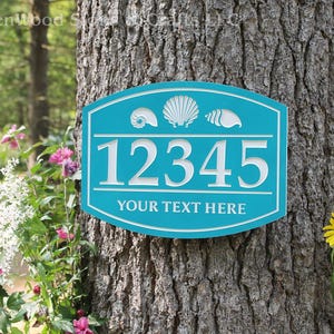 May include: A turquoise blue wooden house number sign with white raised numbers 12345 and the text "YOUR TEXT HERE". The sign has three white seashells on the top.