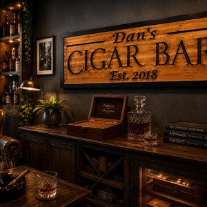 May include: A wooden sign that reads "Dan's CIGAR BAR Est. 2018" hangs above a wooden cabinet. A humidor, decanter, and books are on the cabinet. A leather chair and ashtray are in the foreground. The room has a dark, masculine aesthetic.