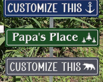 May include: Three rectangular signs mounted on a metal post. The top sign is navy blue with white text reading CUSTOMIZE THIS and an anchor icon. The middle sign is green with white text reading Papa's Place and three pine tree icons. The bottom sign is dark gray with white text reading CUSTOMIZE THIS and a bear icon.