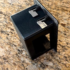 May include: A black, rectangular, open-front box with two silver-colored metal clips attached to the top. The box has a textured surface and is angled towards the viewer. The background is a speckled, light brown surface.