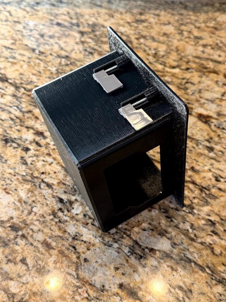 May include: A black, rectangular, open-front box with two silver-colored metal clips attached to the top. The box has a textured surface and is angled towards the viewer. The background is a speckled, light brown surface.