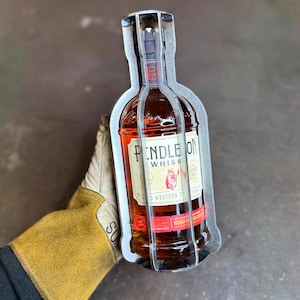 Puede incluir: Una botella de whisky Pendleton, con una etiqueta roja y blanca, se sostiene dentro de un marco de metal. La botella está llena de líquido ámbar. La etiqueta presenta un gráfico de toro y las palabras "Pendleton Whisky".