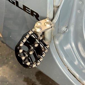 Puede incluir: Peldaño de metal negro con una superficie texturizada y una bisagra de metal plateado unida a una puerta de vehículo gris. La puerta tiene la palabra "Wrangler" impresa en ella.