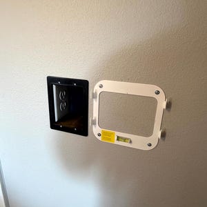 May include: A black electrical outlet box and a white mounting frame with a level are mounted on a light-colored wall. The outlet box has two electrical outlets. The mounting frame is rectangular with rounded corners.