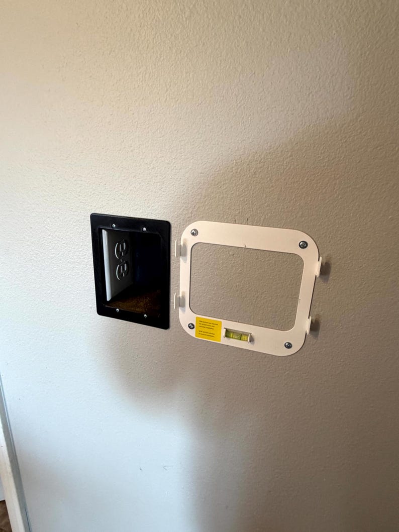 May include: A black electrical outlet box and a white mounting frame with a level are mounted on a light-colored wall. The outlet box has two electrical outlets. The mounting frame is rectangular with rounded corners.