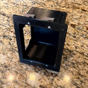 May include: A black, rectangular, 3D-printed box with an open front and a recessed interior. The box has a frame around the opening and metal clips on top. Screws secure the frame. The item is likely a component or housing for another device.