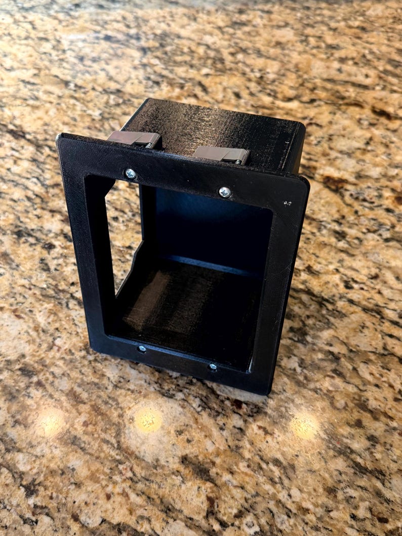 May include: A black, rectangular, 3D-printed box with an open front and a recessed interior. The box has a frame around the opening and metal clips on top. Screws secure the frame. The item is likely a component or housing for another device.