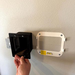 May include: A black rectangular recessed electrical outlet box is being installed on a light-colored wall. A white mounting bracket with a level is positioned nearby. The wall is painted a light beige color.