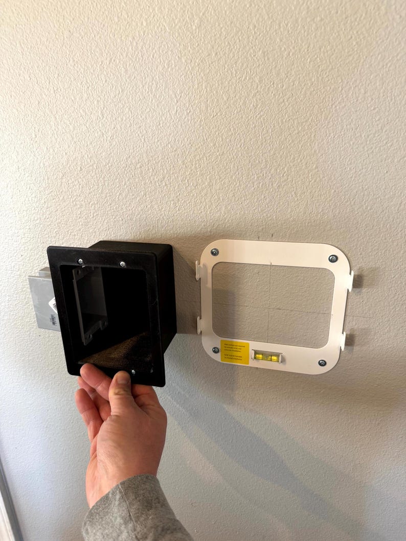 May include: A black rectangular recessed electrical outlet box is being installed on a light-colored wall. A white mounting bracket with a level is positioned nearby. The wall is painted a light beige color.