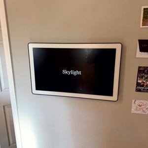 May include: A rectangular digital photo frame with a black screen and a white border is mounted on a light gray wall. The word "Skylight" is displayed in white text on the screen.