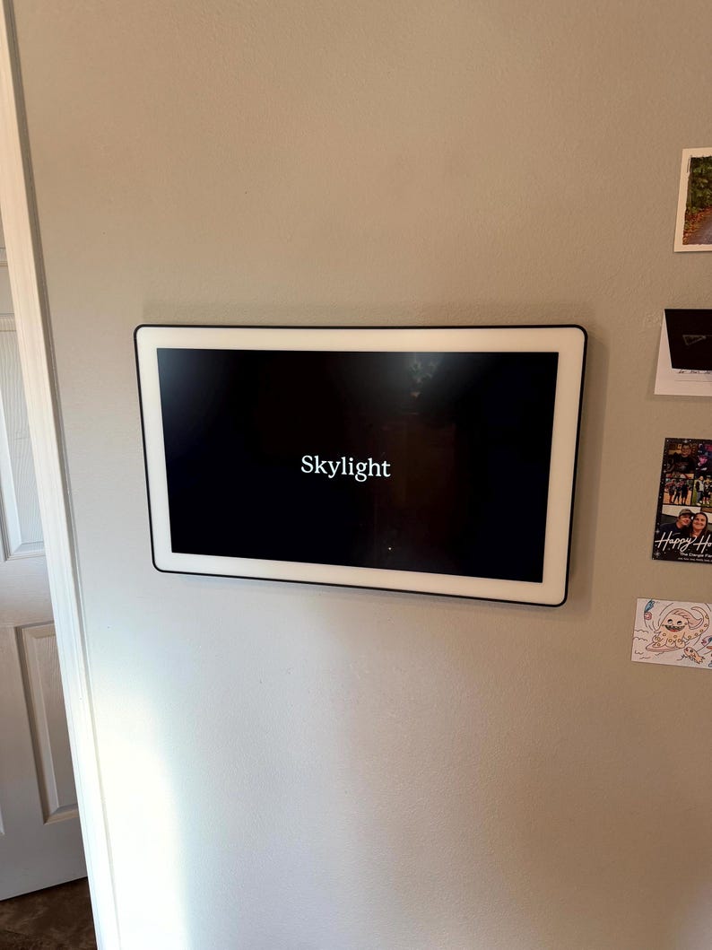 May include: A rectangular digital photo frame with a black screen and a white border is mounted on a light gray wall. The word "Skylight" is displayed in white text on the screen.