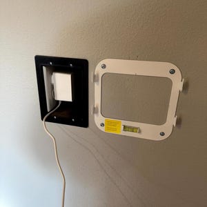 May include: A wall-mounted recessed power outlet with a black frame and a white power adapter. A white mounting bracket with a level is attached to the wall. The bracket has a yellow sticker.