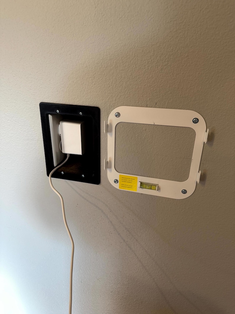 May include: A wall-mounted recessed outlet with a black frame and a white power adapter. A white mounting bracket with a level is attached to the wall. The bracket has a yellow sticker.