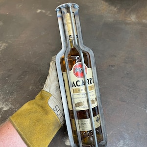 May include: A metal bottle cage with a clear glass bottle of Bacardi Gold rum inside. The bottle cage is made of metal bars welded together to form a cage around the bottle. The bottle is labeled with the Bacardi Gold logo and text. The bottle is sitting on a gray surface.