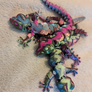 Könnte beinhalten: Zwei bewegliche Eidechsenfiguren mit einem mehrfarbigen Farbverlauf, der zwischen leuchtendem Magenta, Türkis und Limettengrün wechselt. Diese 3D-gedruckten Reptilien haben segmentierte Körper, detaillierte Köpfe und gespreizte Füße.