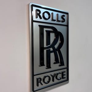 May include: A silver metal sign with the words "Rolls Royce" in black lettering. The Rolls Royce logo is in the center of the sign, with a black "R" inside a double ring.