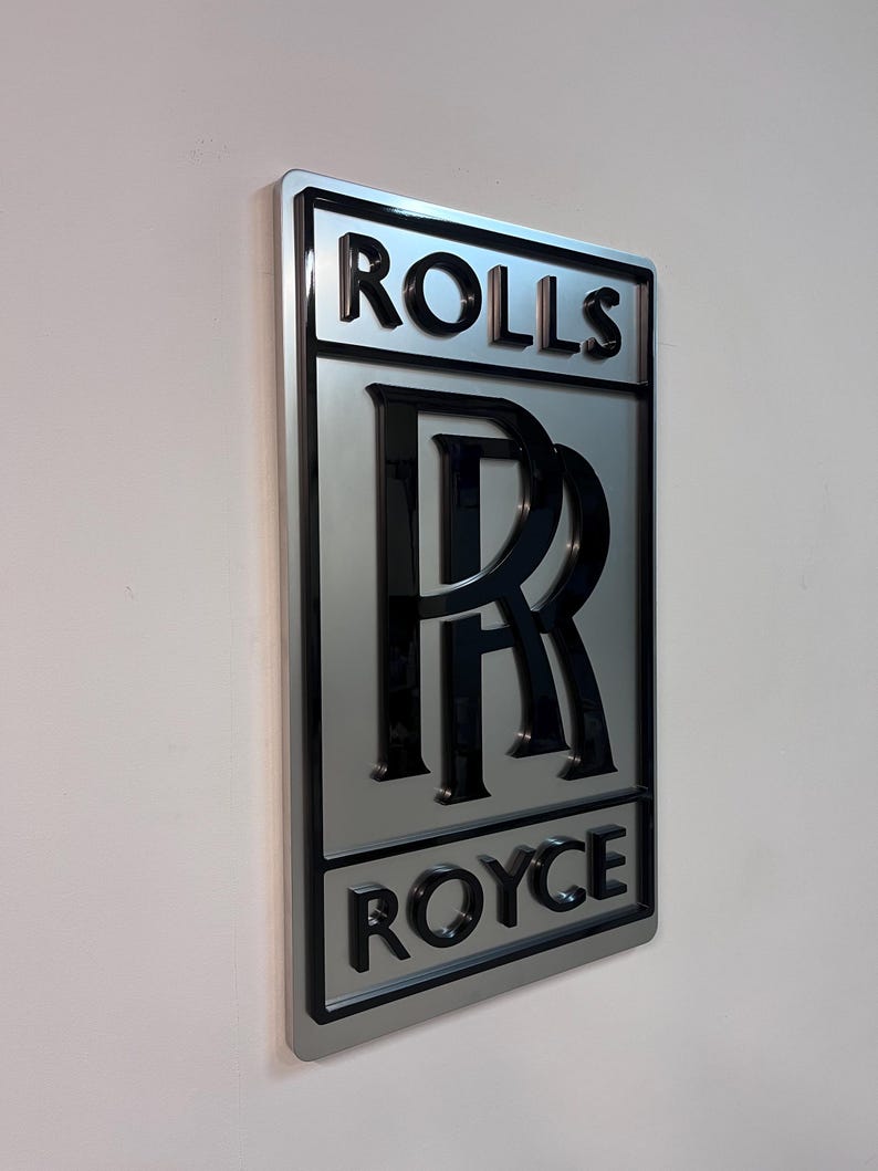 May include: A silver metal sign with the words "Rolls Royce" in black lettering. The Rolls Royce logo is in the center of the sign, with a black "R" inside a double ring.