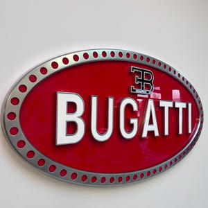 Op de afbeelding: Een rood ovaal bord met een zilveren rand en een wit "BUGATTI" logo. De "B" in het logo is zwart en heeft een witte omtrek.