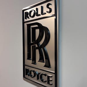 May include: A silver metal sign with a black 3D Rolls Royce logo. The logo features the iconic double R emblem and the words "Rolls" and "Royce" in a black, bold font.