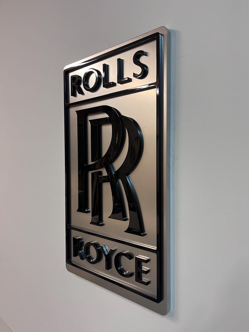 May include: A silver metal sign with a black 3D Rolls Royce logo. The logo features the iconic double R emblem and the words "Rolls" and "Royce" in a black, bold font.