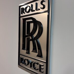 May include: A silver metal sign with a black 3D Rolls Royce logo. The logo features the iconic double R emblem and the words "Rolls" and "Royce" in a black, bold font.