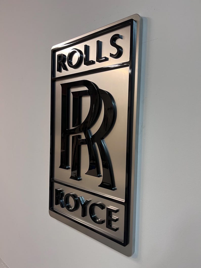 May include: A silver metal sign with a black 3D Rolls Royce logo. The logo features the iconic double R emblem and the words "Rolls" and "Royce" in a black, bold font.