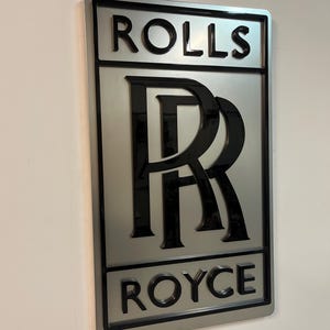 May include: A silver metal sign with a black Rolls Royce logo. The logo features the iconic double 'R' emblem and the words "Rolls" and "Royce" in black lettering.