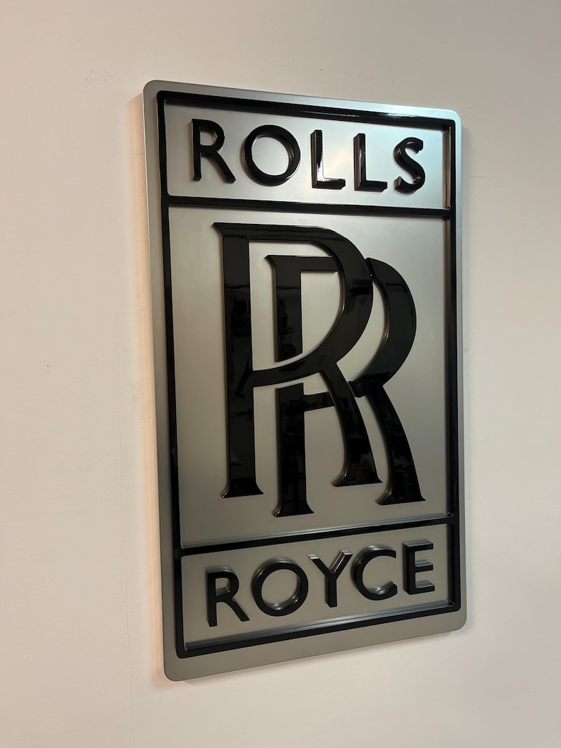 May include: A silver metal sign with a black Rolls Royce logo. The logo features the iconic double 'R' emblem and the words "Rolls" and "Royce" in black lettering.