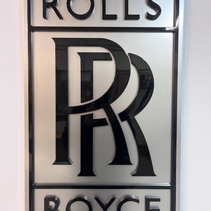 May include: A silver and black 3D sign with the words "ROLLS" and "ROYCE" in black lettering. The Rolls Royce logo is a stylized "R" in black.