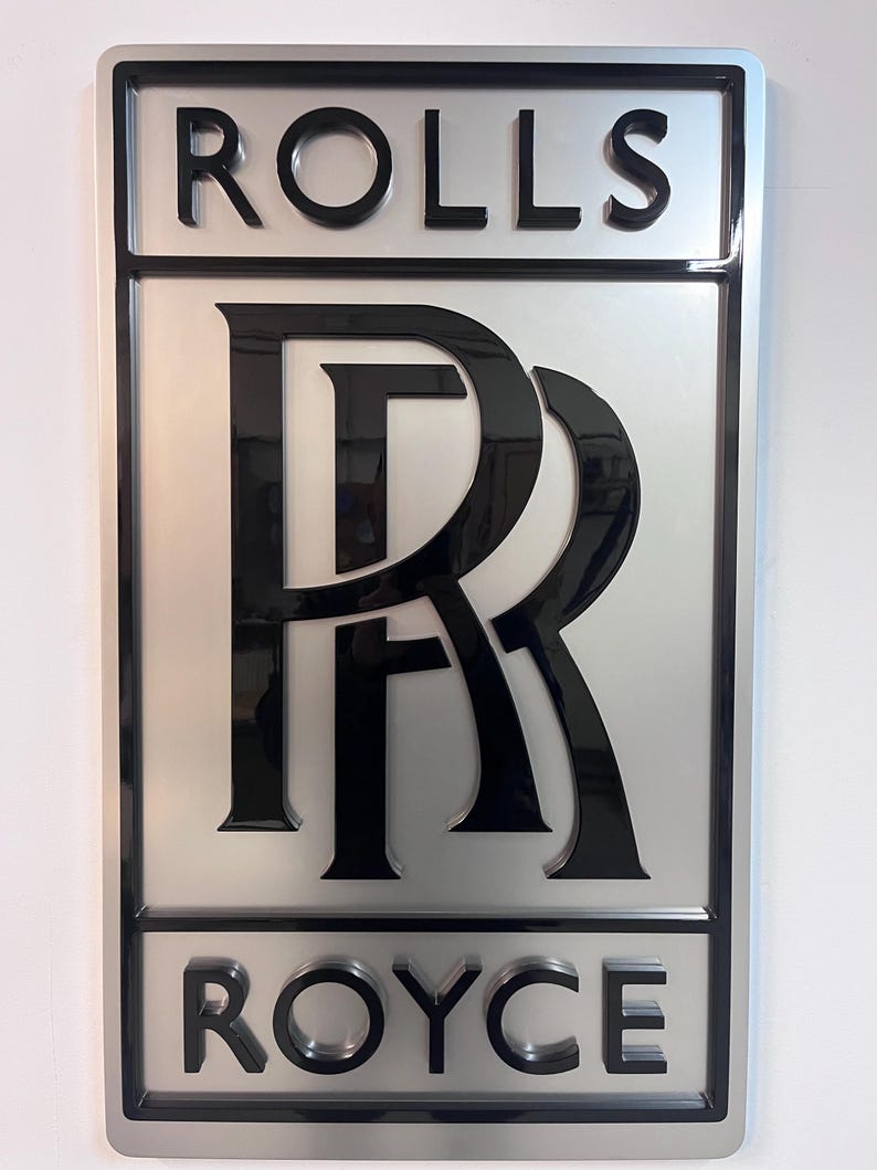 May include: A silver and black 3D sign with the words "ROLLS" and "ROYCE" in black lettering. The Rolls Royce logo is a stylized "R" in black.